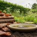 Ground-level view of a wildlife garden with a log pile, leaf litter, a shallow water dish, and native hedge vegetation supporting insects and small wildlife.